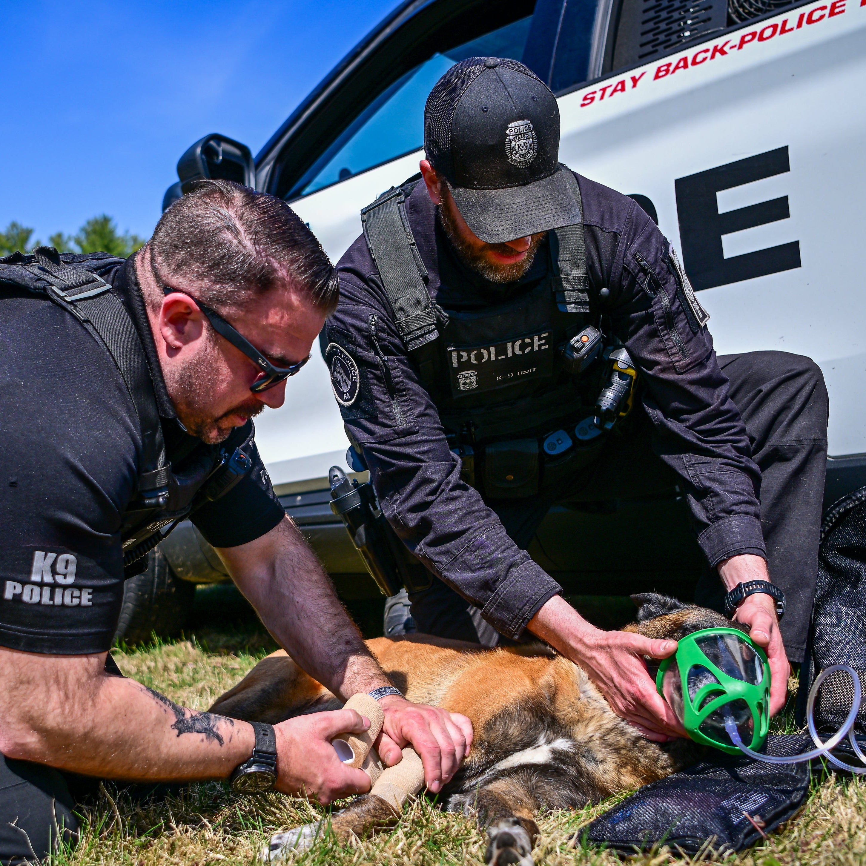 Two police officers attending to a dog near a police vehicle with 'Stay Back Police' written on it.